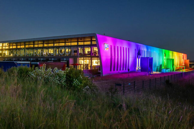 Ein großes Produktionsgebäude ist zu sehen. Es ist Abend und das Gebäude wird mit Regenbogenfarben angestrahlt. 