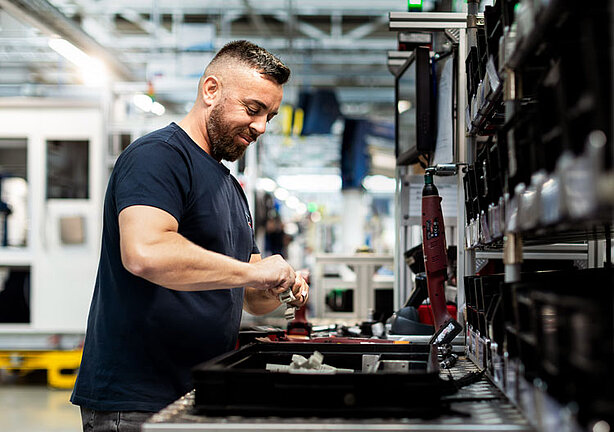 Ein Mann arbeitet an einer Werkbank in einer industriellen Umgebung. Sie hantiert mit Werkzeugen und Ausrüstung. Im Hintergrund sind Maschinen und andere industrielle Elemente zu sehen.