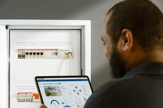 Person nutzt ein Tablet mit Energie-Management-Software vor einem geöffneten Sicherungskasten mit Schaltern und angeschlossenem Netzwerkkabel.