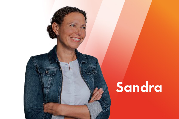 Portrait einer dunkelhaarigen Frau Frau, die eine weiße Bluse und eine Jeansjacke trägt. Sie hat lachend die Arme vor der Brust verschränkt und schaut nach rechts.  