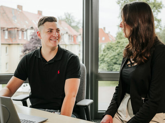 Ein Mann und eine Frau sitzen an einem Schreibtisch und schauen sich lachend an. 
