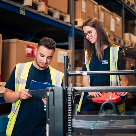 Vor einen Lagerregal steht eine Auszubildende auf einem Stapler neben einem anderen Auszubildenden. Beide schauen auf ein Klemmbrett. 