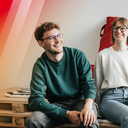 Ein Mann und eine Frau sitzen lachend auf Paletten-Möbeln. 