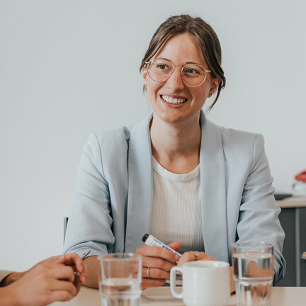 Eine Frau mit dunklen Haaren und Brille sitzt an einem Tisch. Auf dem Tisch sieht man Gläser, Stifte und Karteikarten.