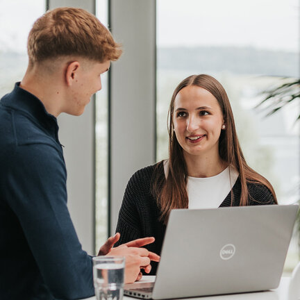 Zwei junge Erwachsene sprechen miteinander. Vor ihnen steht ein aufgeklappter Laptop.