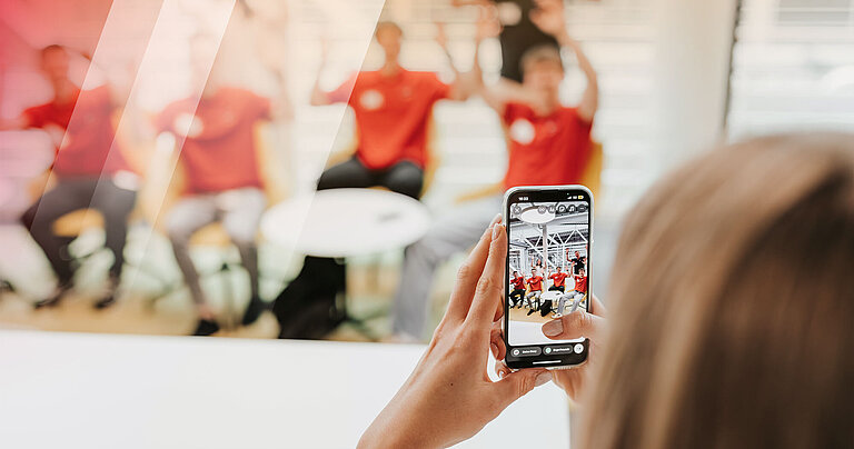Im Vordergrund ist ein Handy, mit dem sechs Personen im Hintergrund fotografiert werden.