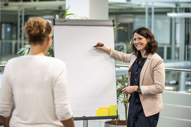 Man sieht zwei Personen. Eine Frau steht an einem Whiteboard zur Kamera gewendet und schreibt dort gerade etwas. Eine zweit Frau steht mit dem Rücken zur Kamera und schaut auf das Whiteboard. 