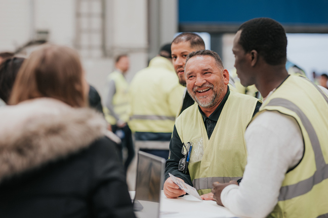 Mehrere Personen, die Warnwesten tragen. Der Fokus liegt auf einem lachenden Mann, der an einem Stehtisch steht und eine Frau anschaut. 