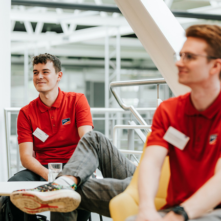 Zwei junge Männer in roten Poloshirts sind zu sehen, wobei die Person im Vordergrund unscharf fotografiert ist. 