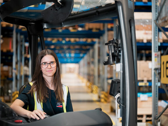 Eine junge Frau mit dunklen Haaren und Brille sitzt in einem Stapler in einer Logistikhalle. Sie trägt eine Warnweste und schaut freundlich in die Kamera.