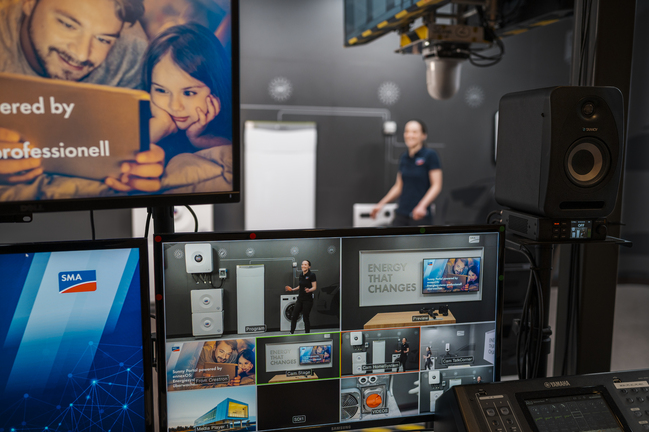 Mehrere Monitore und technische Geräte in einem Studio-Setup, zeigen verschiedene Video-Feeds und Präsentationsinhalte zu Energie- und Technologieprodukten, darunter SMA-Branding und der Text ‚Energy that changes