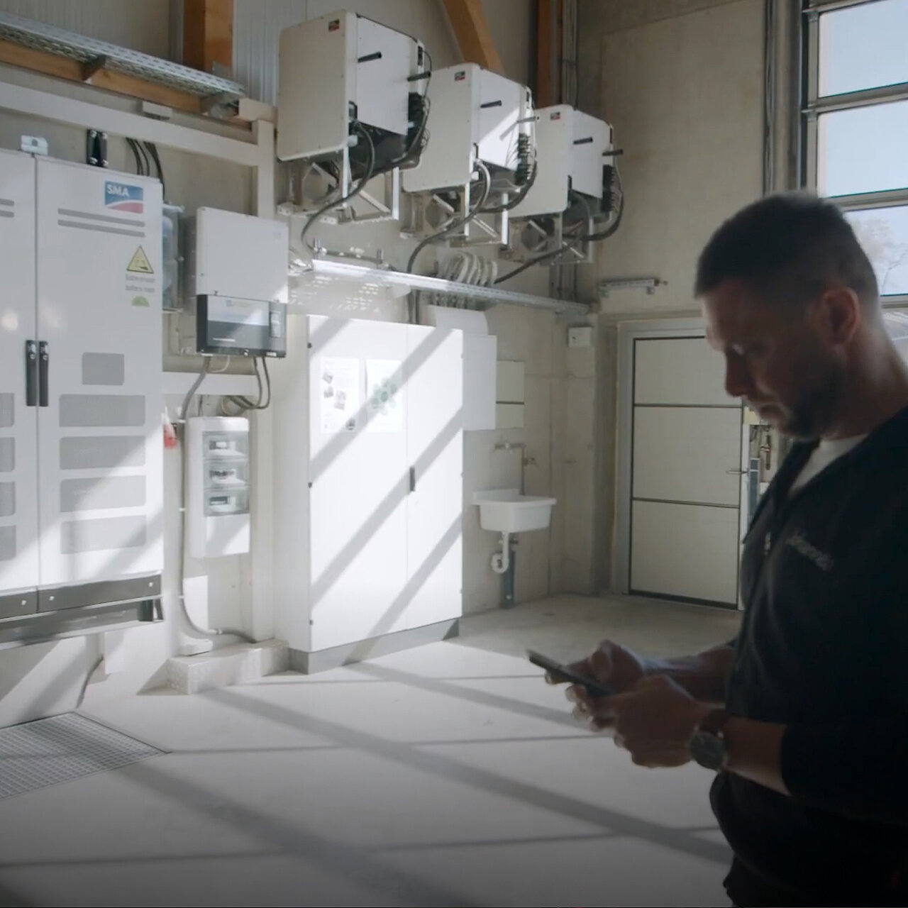 Eine große Halle in der mehrere SMA Sunny Tripower CORE1 an der Wand hängen, ein SMA Schaltschrank, sowie ein weiterer Sicherungsschrank, davor steht ein Mann, der auf seinem Mobiltelefon schaut.