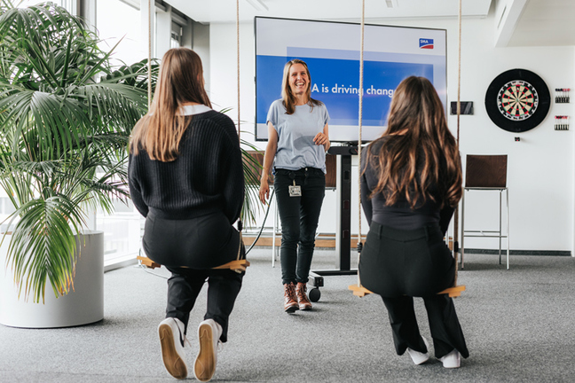 Drei Personen in einem modernen Büro. Zwei sitzen auf Schaukeln, eine spricht vor ihnen. Im Hintergrund ein Bildschirm mit "SMA is driving change". Links eine Pflanze, rechts ein Dartboard.