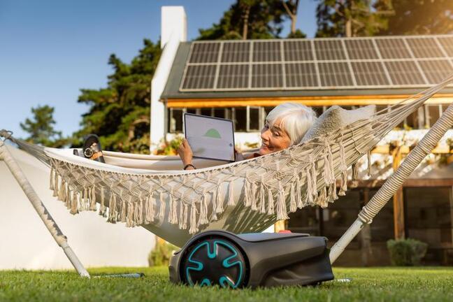Person using a laptop in a hammock in a garden, with a robotic lawn mower in the foreground and a house with rooftop solar panels in the background.