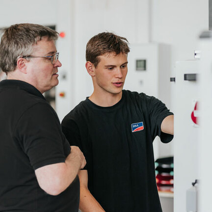 Ein Ausbilder und ein Auszubildender stehen vor einem Schaltschrank und sehen sich gemeinsam etwas an.