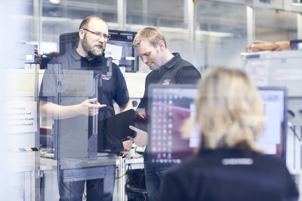 Drei Personen sind in einer IT-Umgebung zu sehen. Man sieht im Hintergrund Datenspeicher und Computer. 