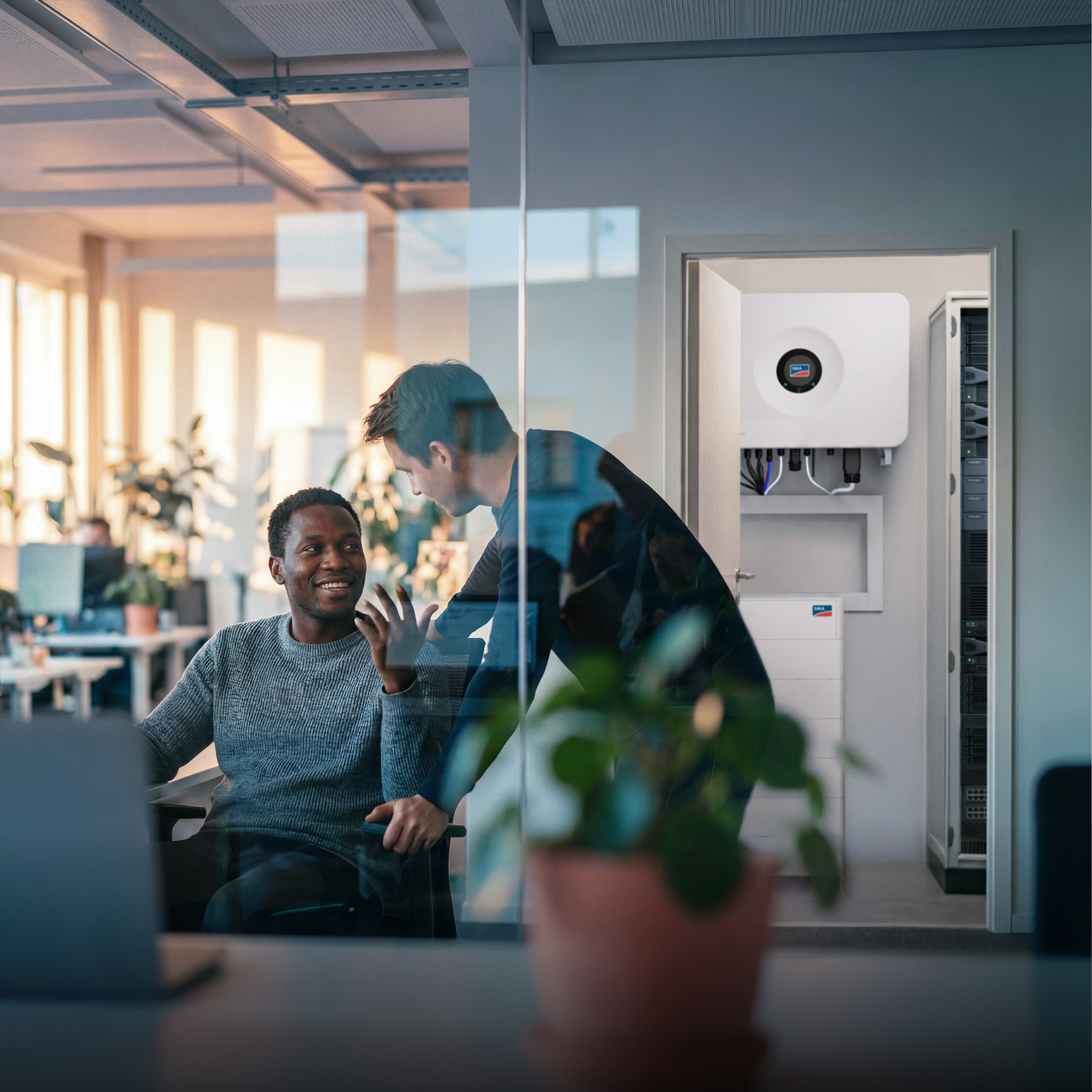 Two men are talking in an open-plan office; behind them, the door to a distribution room is open, and inside stands an SMA Storage M, with a Sunny Tripower Hybrid X mounted on the wall.