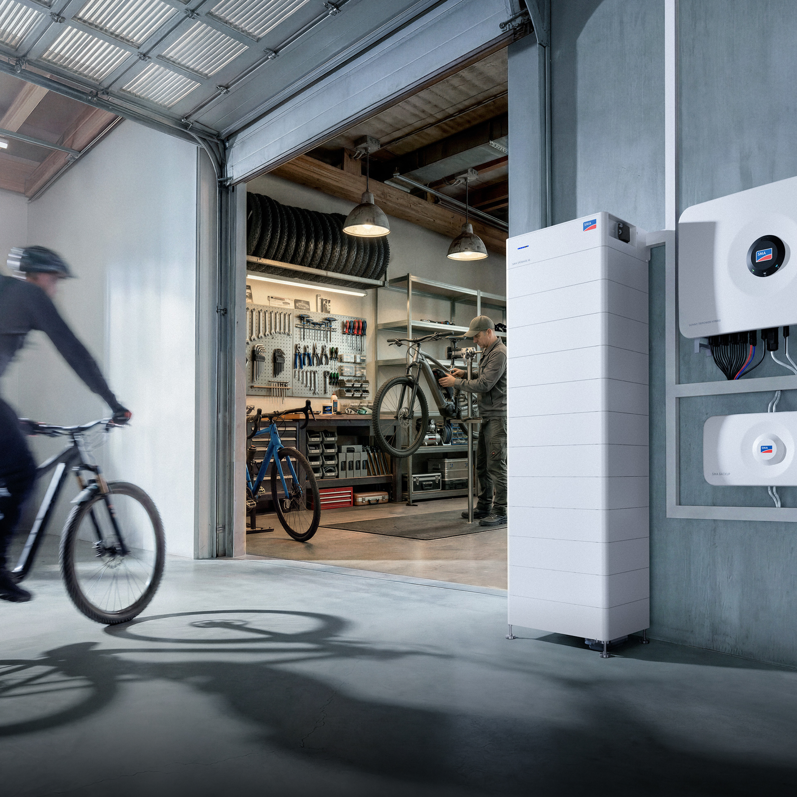 A bike repair shop in a warehouse; in front of it stands an SMA Storage M battery storage system; on the wall hang a Sunny Tripower Hybrid X inverter and an SMA Backup.